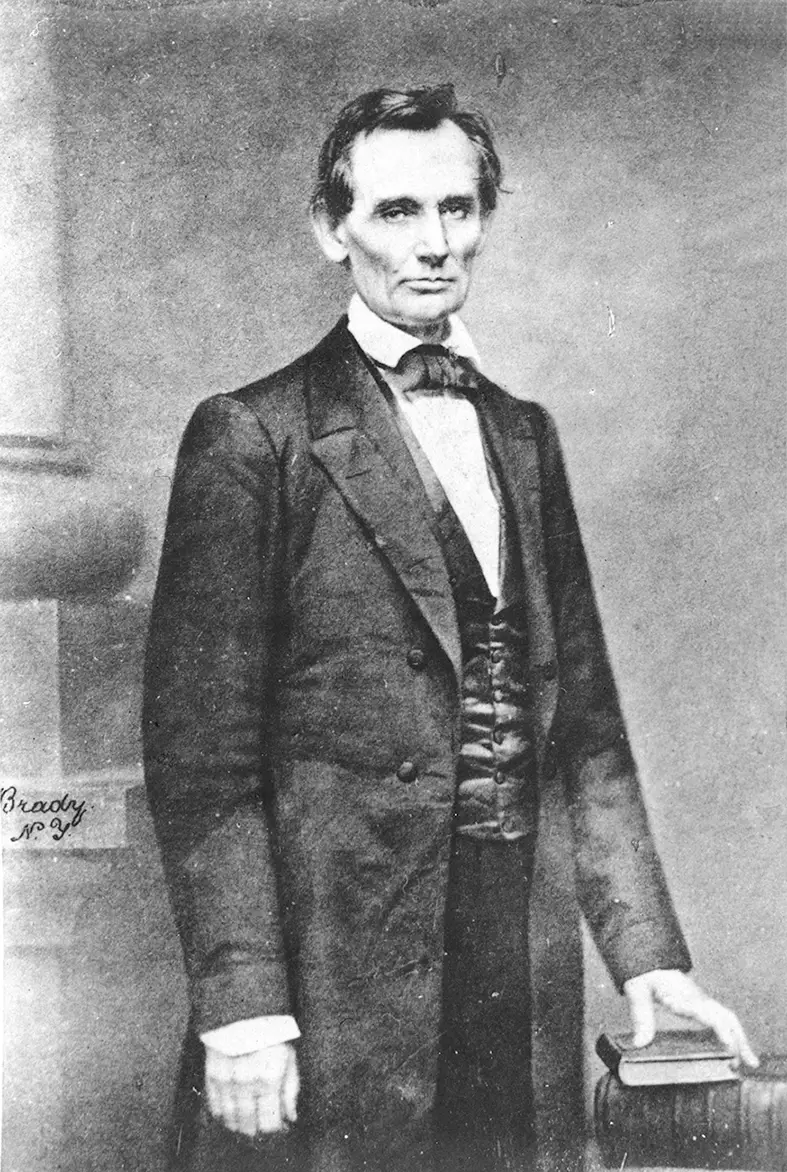 Black and white photo of Lincoln posing for a portrait, dressed in a suit, with his hand on a book, looking straight into the camera.