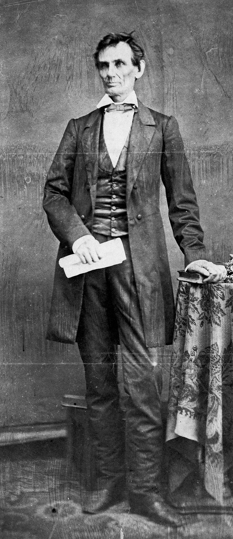 Black and white photo of Lincoln, posed with a scroll in one hand, the other resting on a book on a table.