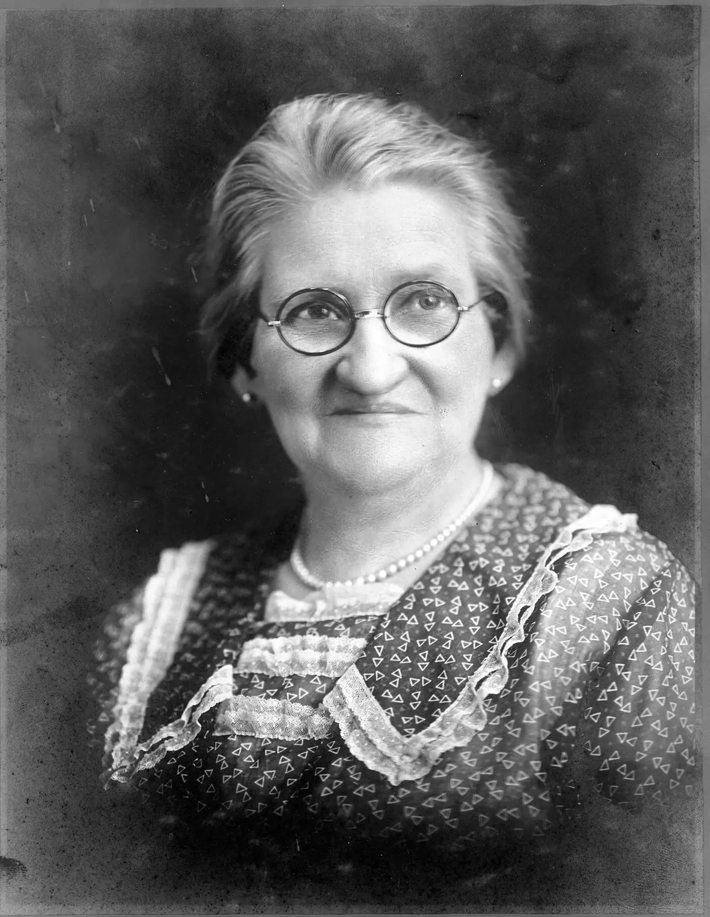 Black and white portrait of Augusta Mau, a middle aged woman in a triangle-patterned dress with lace trim, necklace, and wearing round glasses.