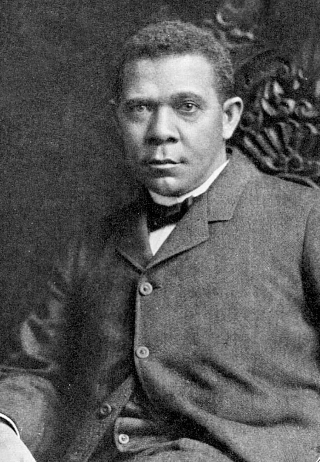Black and white photo of a Black man sitting in a chair. He is wearing a suit with bowtie
