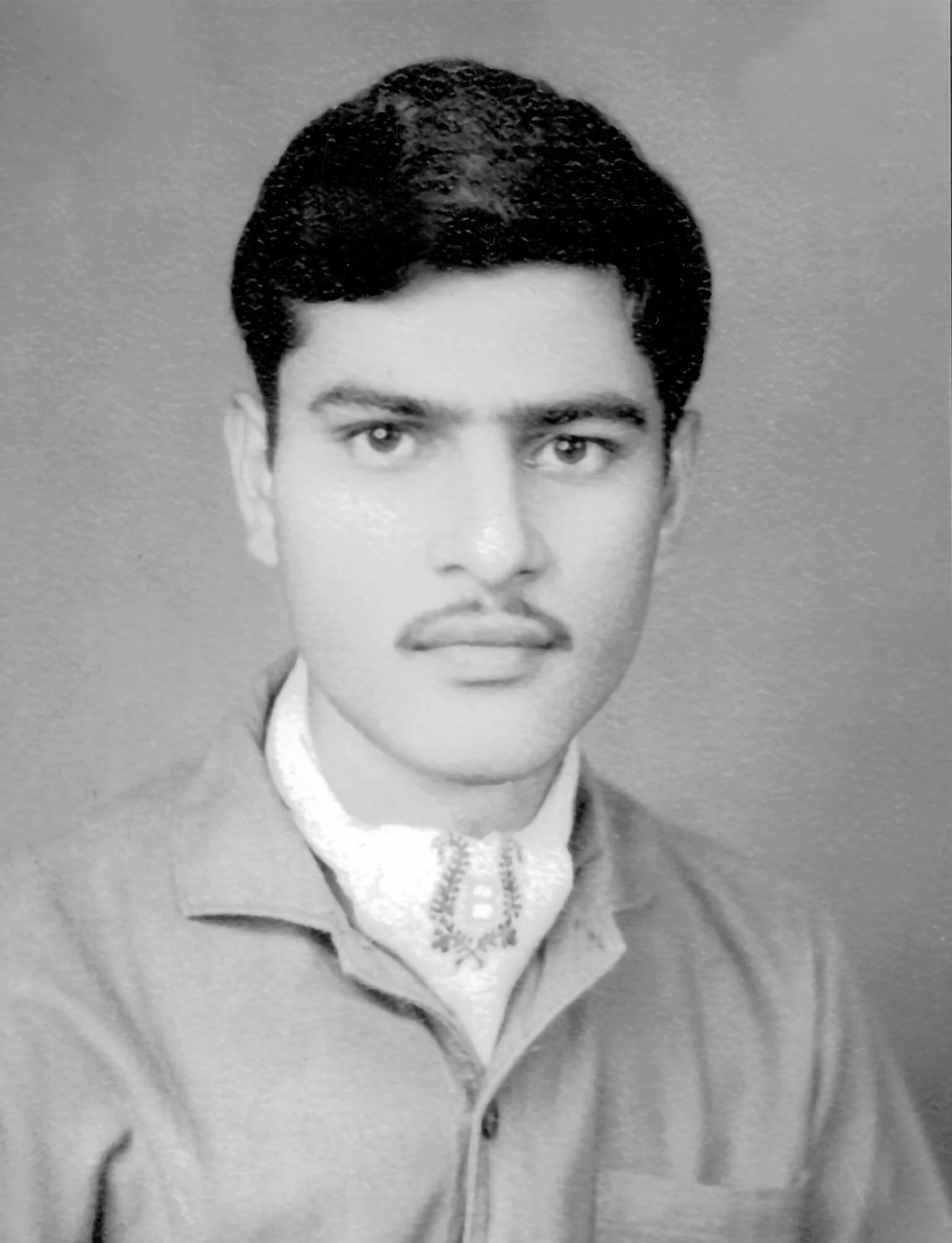Black and white photo of Balwant Singh, with a faint mustasche, black hair, and wearing a light-colored collared shirt and scarf.
