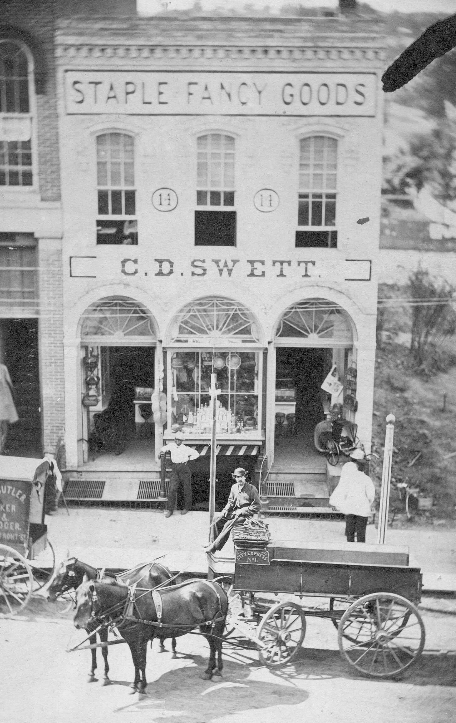 A man sitting in a carriage pulled by two horses is on the street outside of a two story brick building. The building has three large arched windows for the storefront on the first floor.