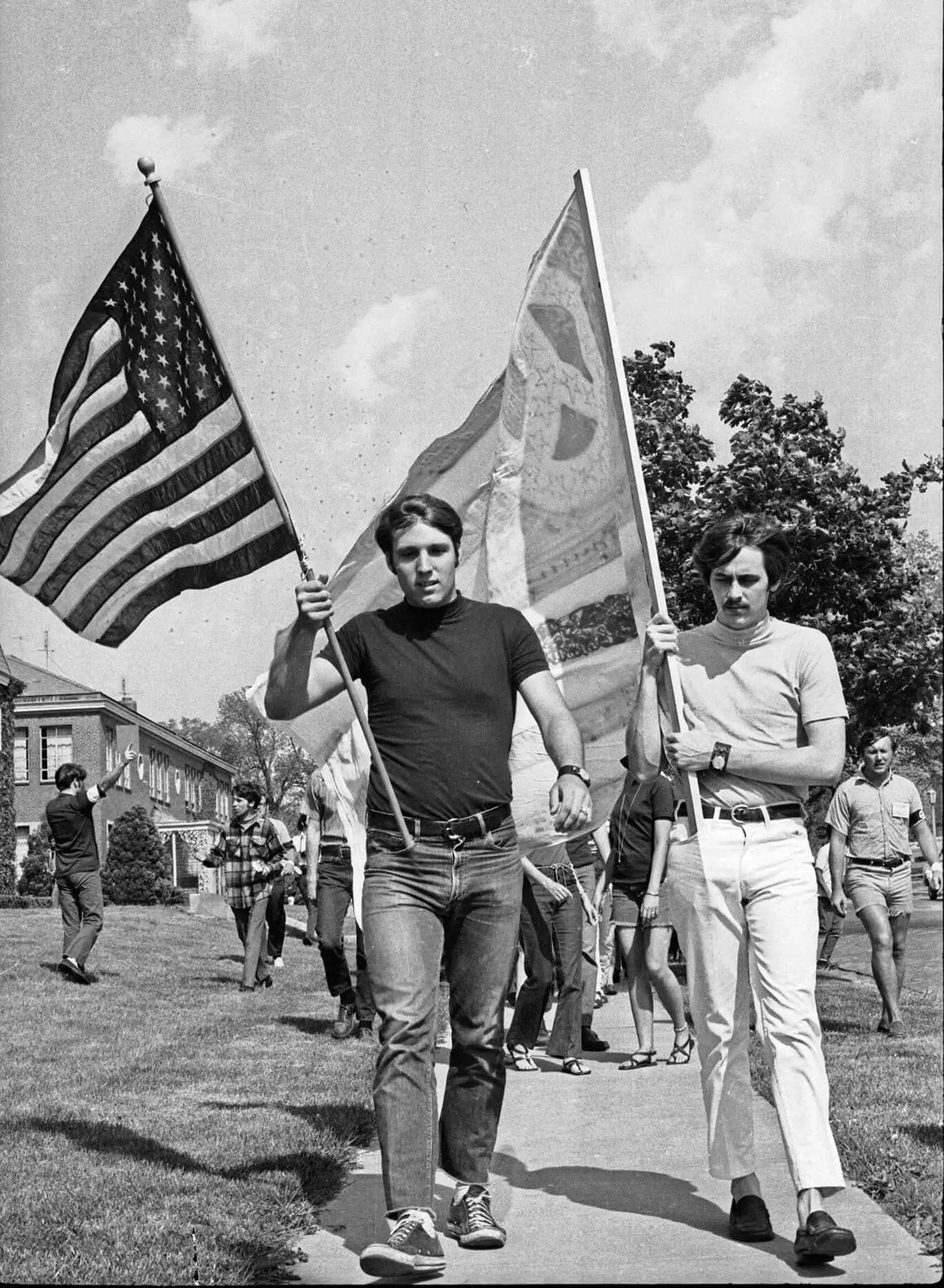 Two white men are walking and holding flags. One man with dark hair, a dark shirt, and dark jeans holds the American flag. A man next to him with dark hair and a mustache is wearing a light colored turtle neck and light colored pants. He is holding a hand-drawn peace flag.