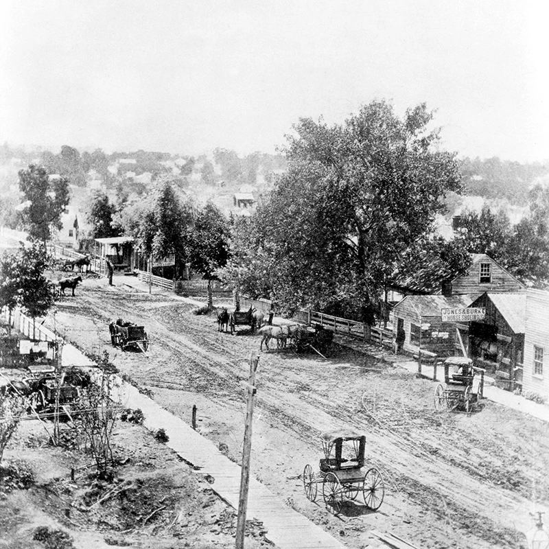 Black and white photo of a dirt street populated with horses and horse-drawn carriages. There are deep ruts in the street from the carriage wheels. The sidewalks are wood planks. In the center foreground of the image is one pole, used to carry telegraph wires.