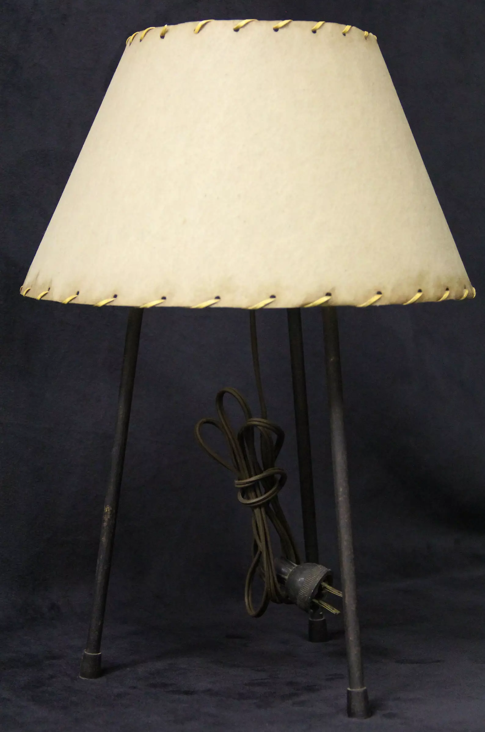 Photo of a lamp with 3 dark legs, a cord, and a white lampshade that has a thin leather strip weaved around both the top and bottom of the shade.
