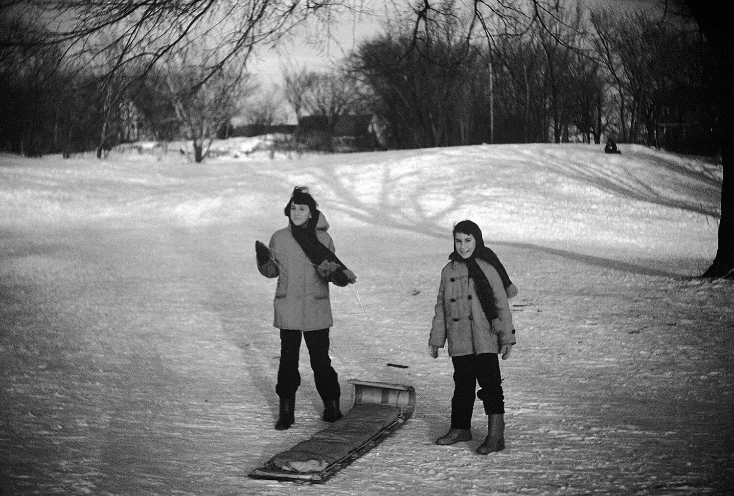 Portrait of two children dressed in winter gear, standing in an open field with a toboggan.