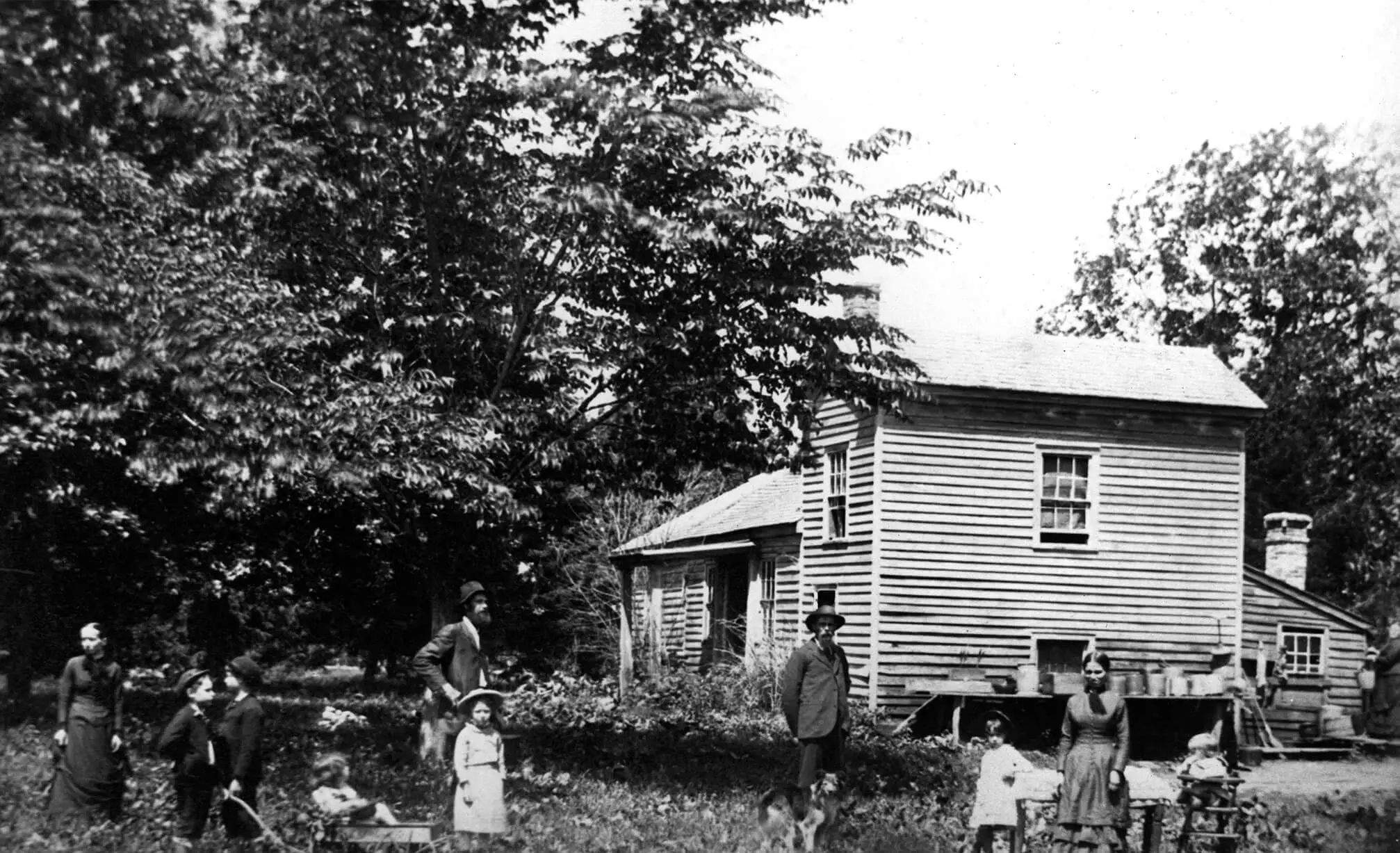 Photo of a light-colored two-story house. There are many trees surrounding the house. Several children, men and women, stand in front