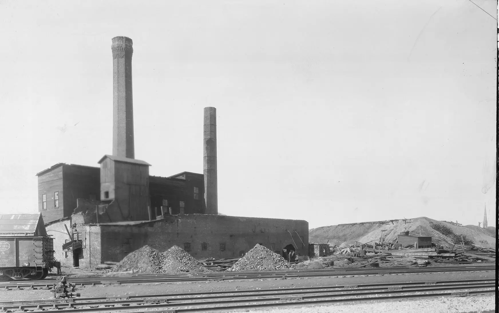 Black and white illustration of a brick building with two large smokestacks. A train car can be seen on the bottom left of the photo with a pile of rubble, likely coal, on the right side.