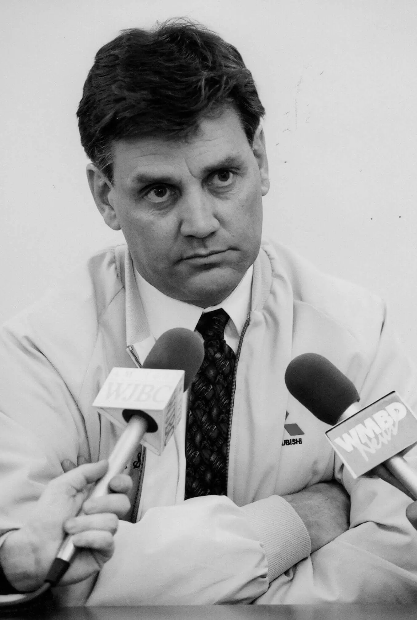 White man with two microphones held up to him. He looks unhappy. He is wearing a mitusbishi zip-up jacket, white shirt and tie. He has dark short hair.