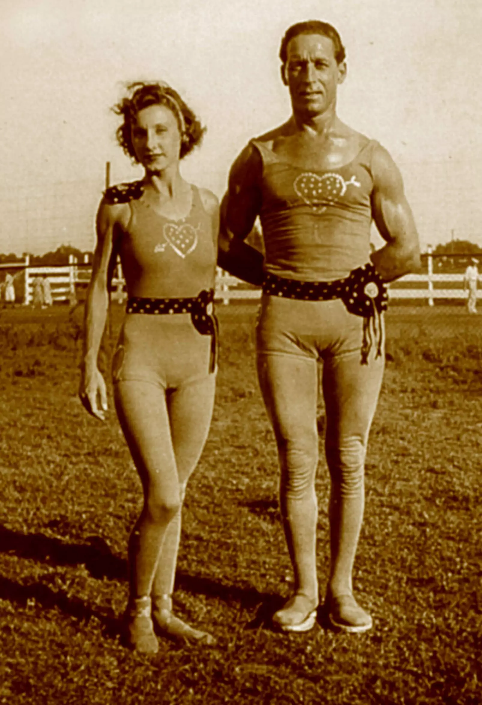 They are standing outdoors in the grass. They are wearing matching outfits, skin tight leotard with a tank top style at the top, shorts at the bottom. On their chest is a heart design with an arrow through the middle. They have a decorative ribbon belt around their waists, and tights cover their entire legs and feet.