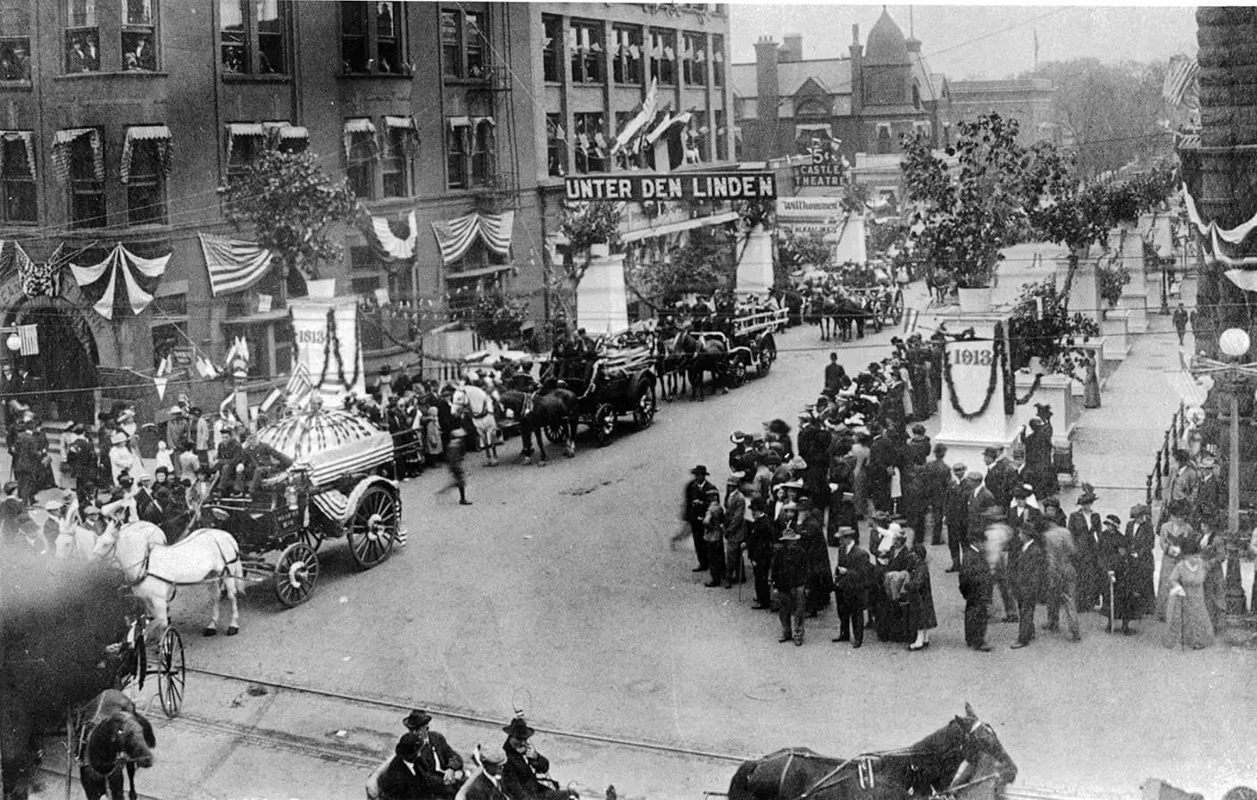 Photo of a large crowd lining the street, watching a parade of horsedrawn wagons decorated as floats. There is bunting displayed on some of the buildings along the parade route.
