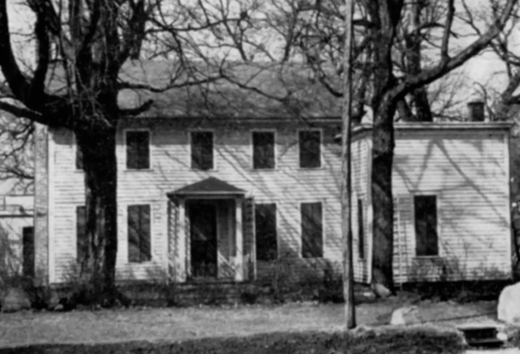 A black and white photograph of a two-story wooden frame house with two trees in the yard.