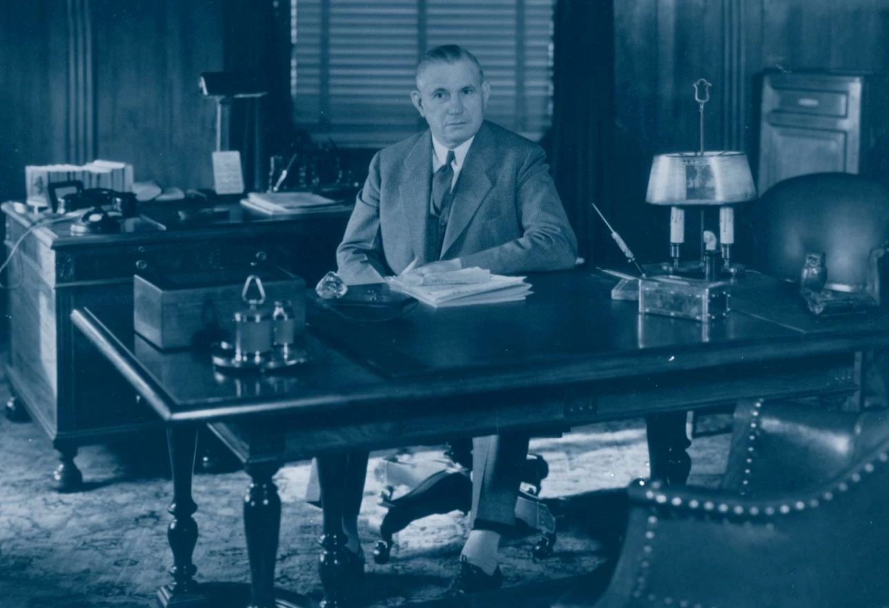 Man with hair combed backwards wearing a suit and tie sits at a desk. His left elbow is resting on the desk. It has a decorative carpeting, leather chairs, lamps, a phone, an ink pen, ink blotter, and some papers. The office has wood paneling on the walls.