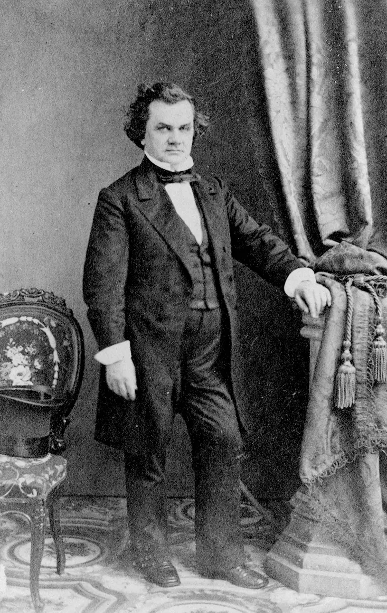 Black and white full-length photograph of short white man in suit, posed in front of curtain and chair.
