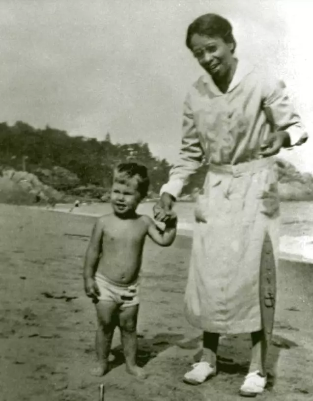African American woman wearing a uniform and apron holds the hand of a small white boy. he is only wearing underwear or a bathing suit. They are on the beach, the water and coastline are seen in the background.