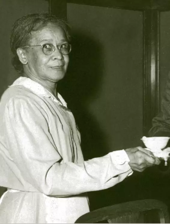 An African American woman wearing a uniform holds a teacup on a saucer and looks to her right, towards the camera. She seems to be possibly taken by surprise by the photo being taken.