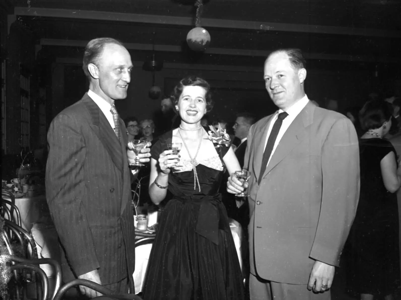 Black and white photo of Proctor Brown and a couple composed of a tall balding man in a suit and woman with parted hair in a dark dress, conversing at a party.