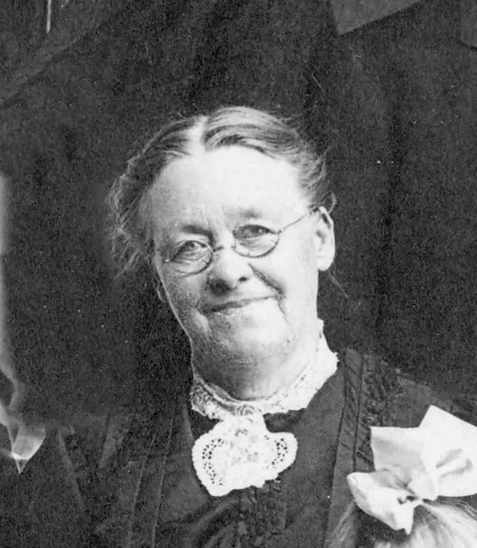 Black and white photo of a white woman. She is wearing a pair of round wire spectacles and a black dress with lace trim. She's smiling subtly and tilting her head. Her hair is parted down the middle and pulled back.