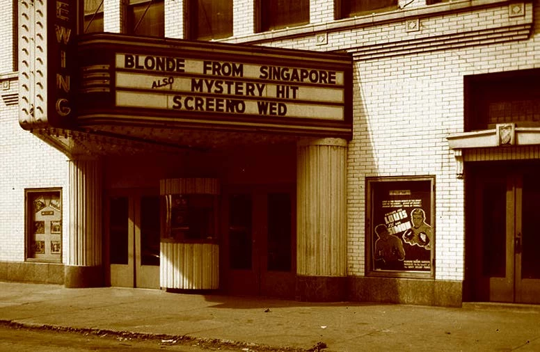 Black and white photo of the front of a theater, with a marquee sign that projects in a large triangle shape over the sidewalk. The sign has removable lettering, announcing what movies are playing.