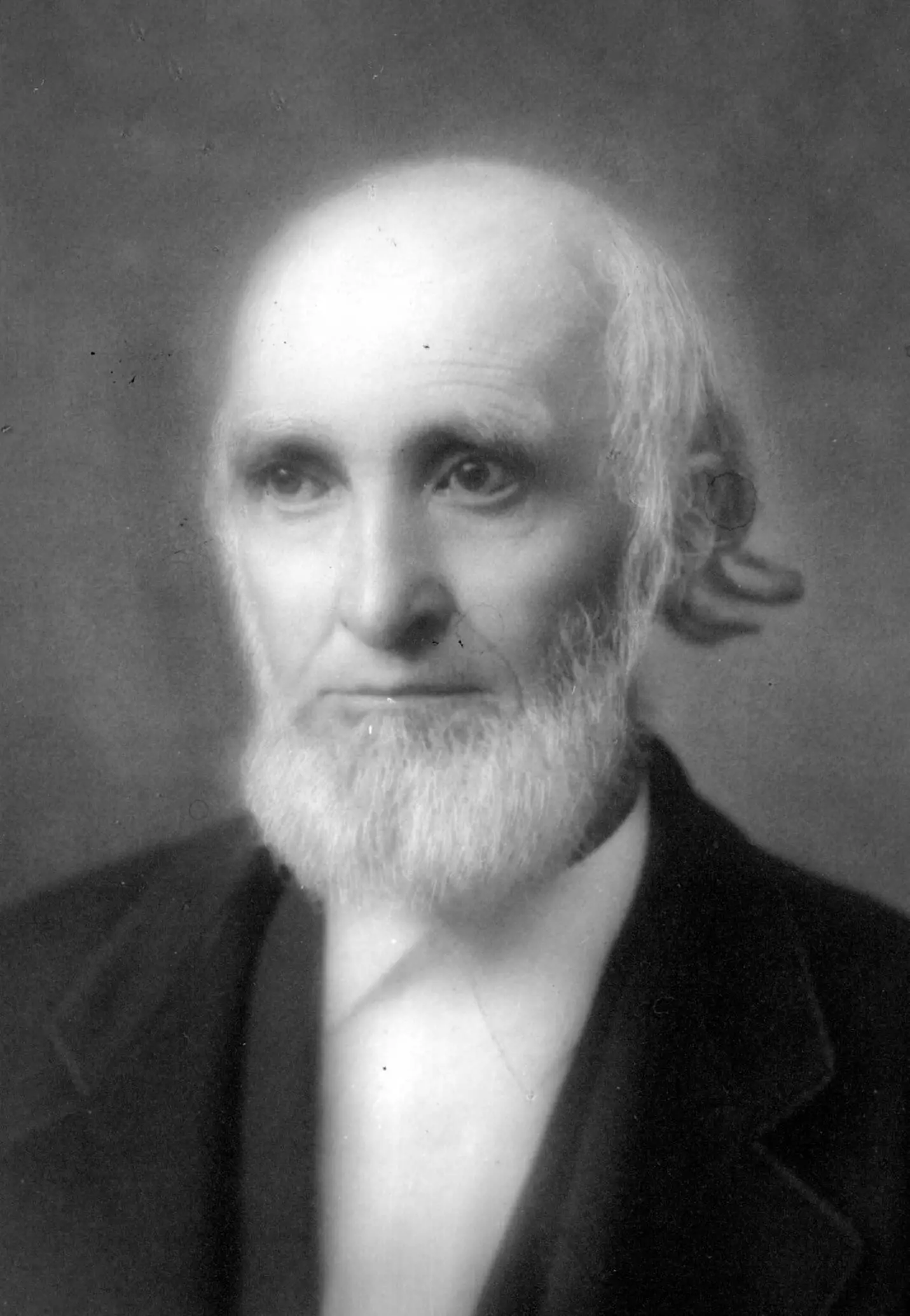 A portrait of a white man with a short white beard wearing a dark coat and a white shirt.