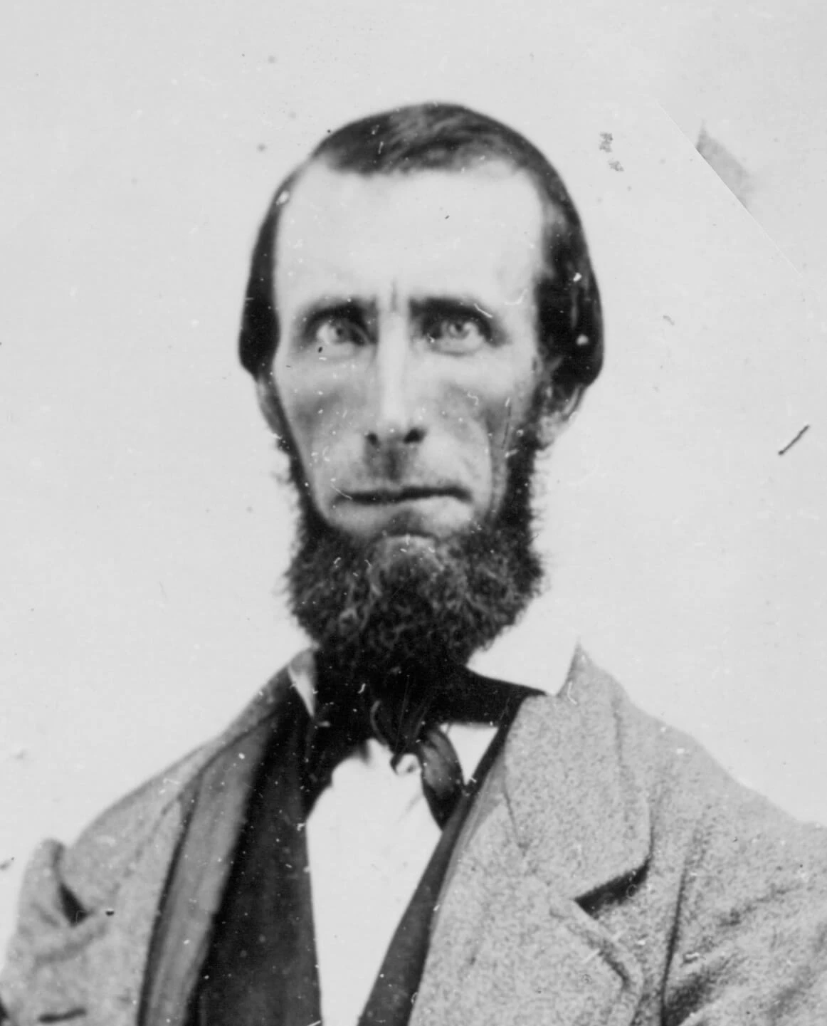 Portrait of a gaunt-faced man with a full beard & sideburns along with a receding hairline wearing a suit and tie.