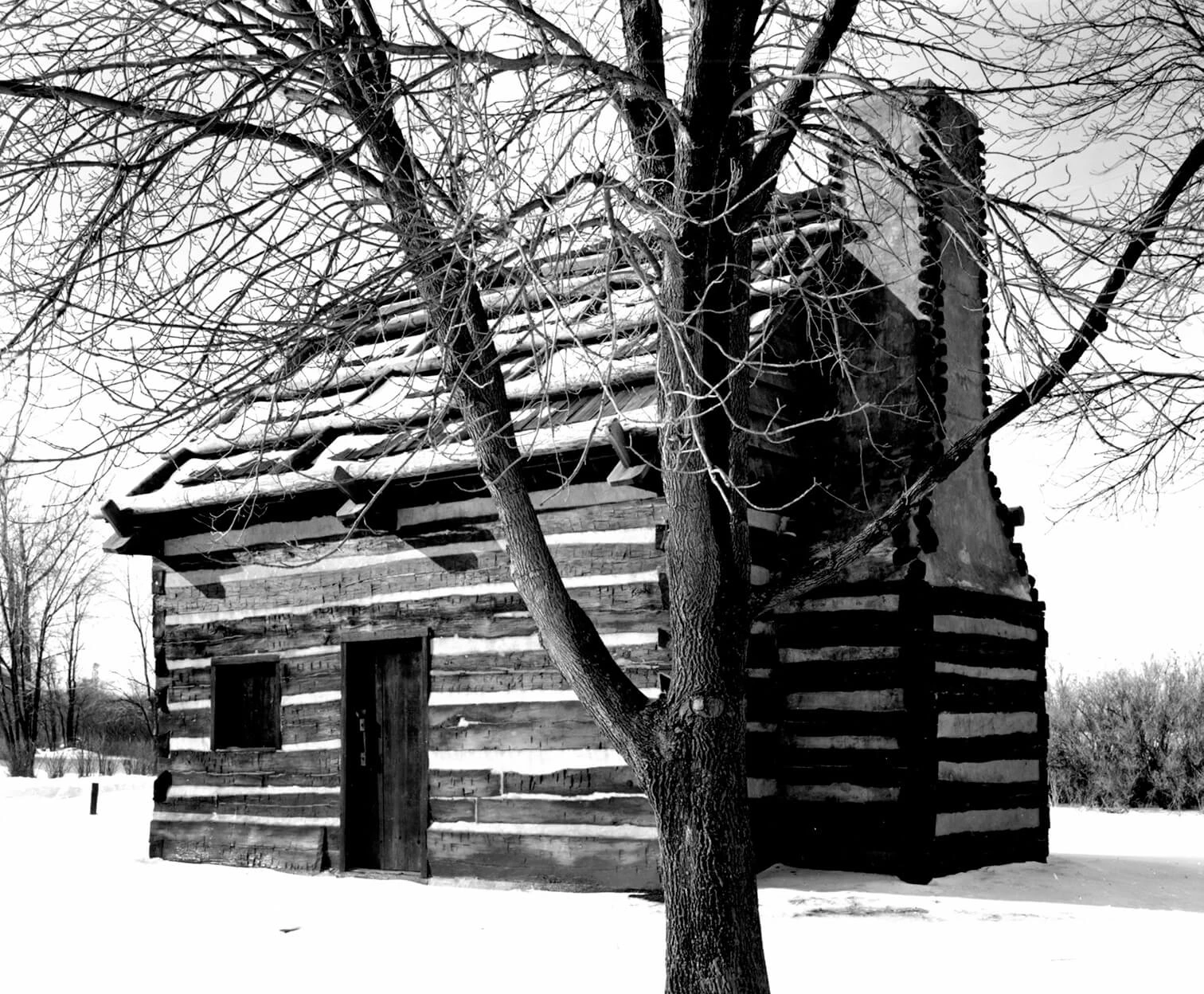 Black and white photo of a rectangular log cabin with a door and window on the longer side and chimney on the shorter side. There is a large tree in the foreground next to the cabin and snow on the ground.