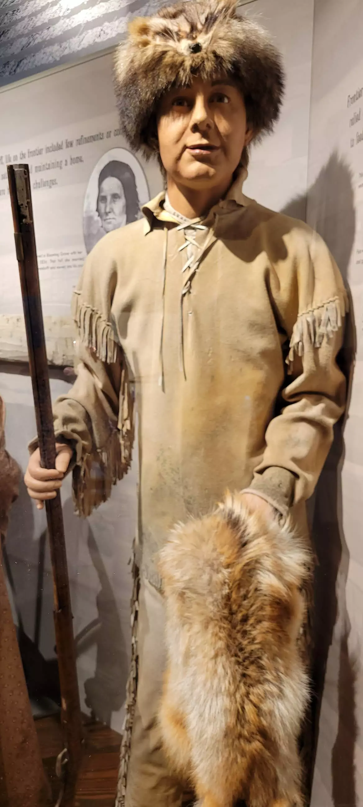 A white, male mannequin dressed in buckskin, standing with a wooden rifle in one hand and a fur in the other hand.