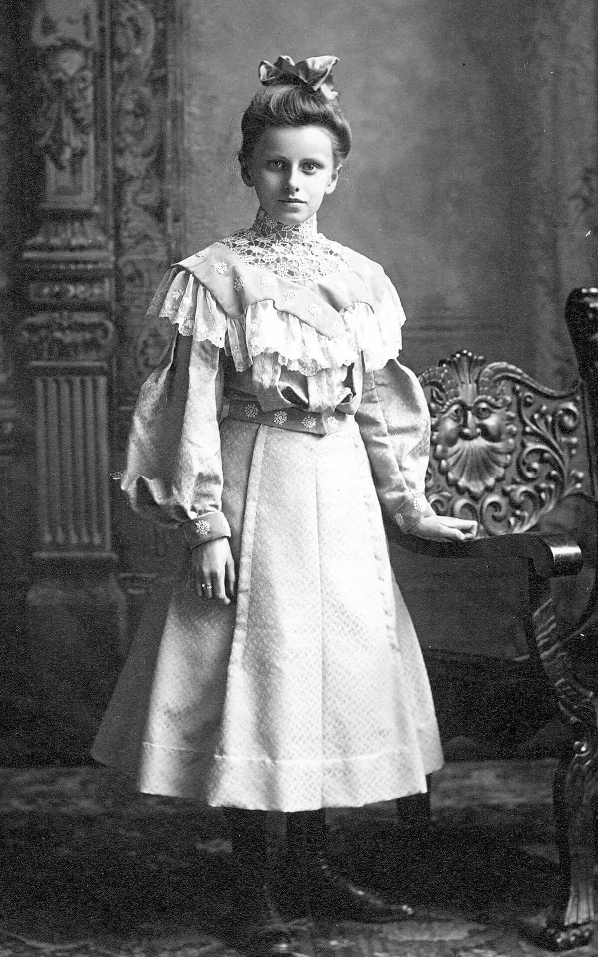 Portrait of Katie Mau, wearing a lightly patterned floral and lace dress, with her hand resting on a chair that features a bearded face carved into it. Her dress is shin-length and she is wearing dark boots and stockings.