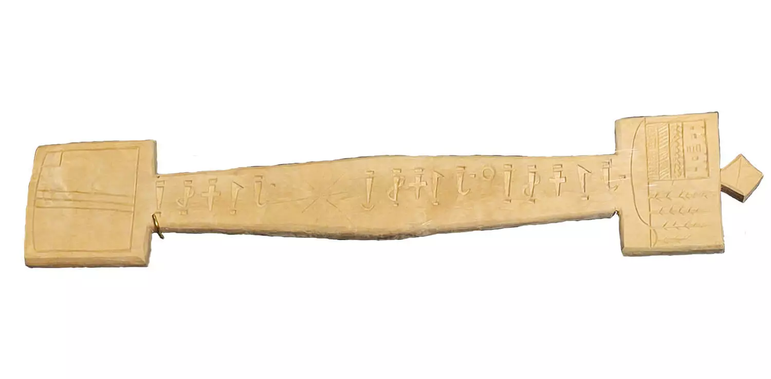 A flat, long wooden prayer stick. The middle is slightly wider, then tapers towards the ends, and on each end there is a rectangle. The whole length of the prayer stick has symbols carved into it.