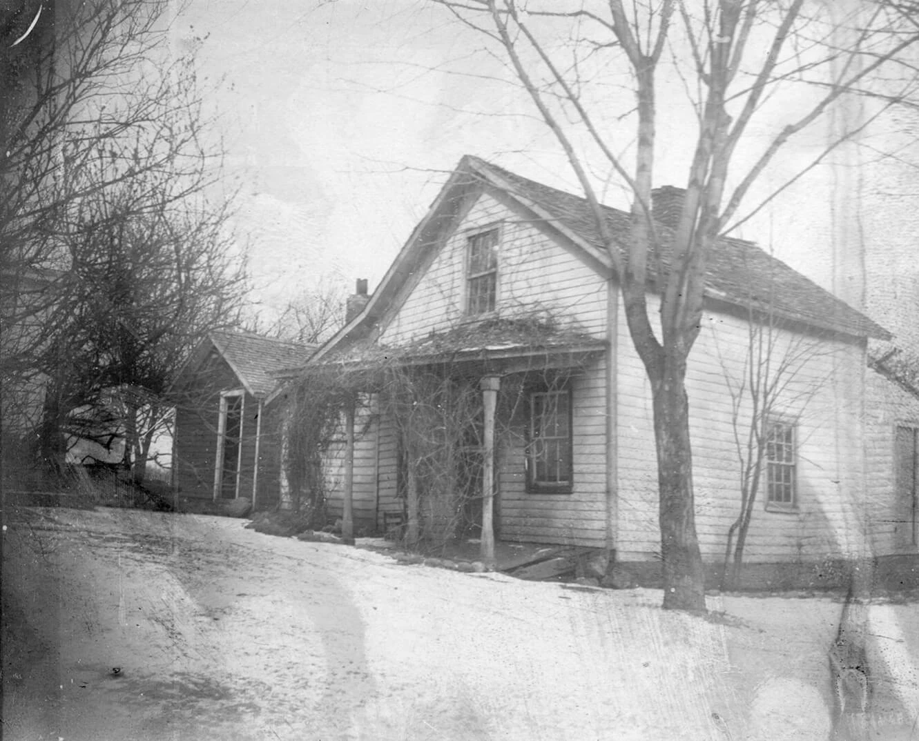 Black and white photo of the Horatio Petit house. The porch is covered in vines, and a tree stands in the front corner of the yard. It appears to have been taken in winter, with snow on the ground.