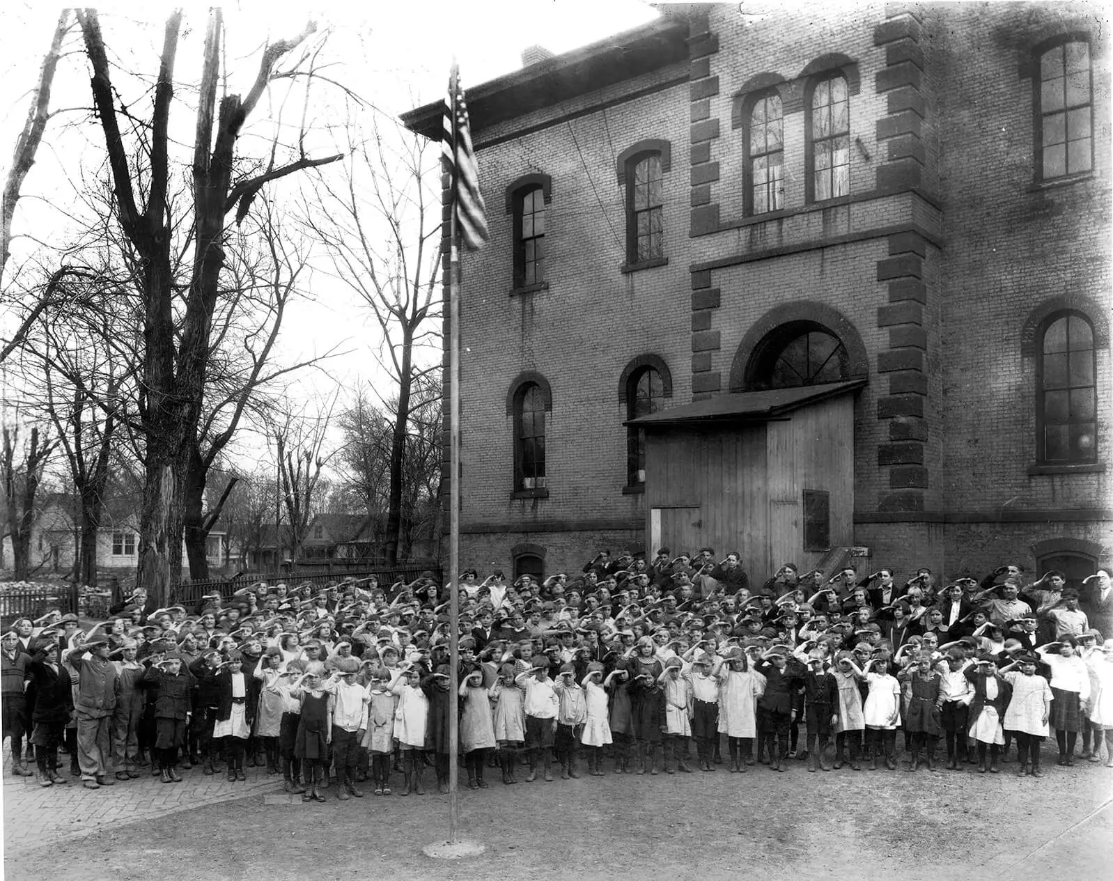 Black and white photo of over 100 schoolchildren touching their hats outside an elementary school.