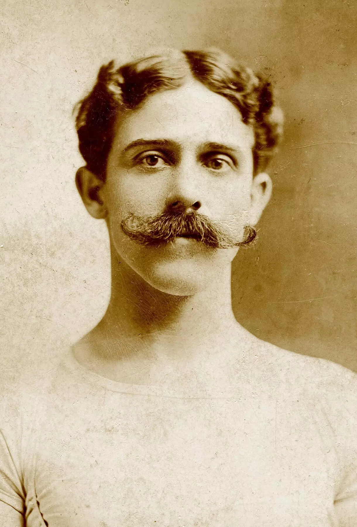 Black and white portrait of a light-skinned man with wavy dark hair parted down the middle, and a large handlebar mustache. He is wearing a very tight white shirt with no collar, possibly stretchy material.