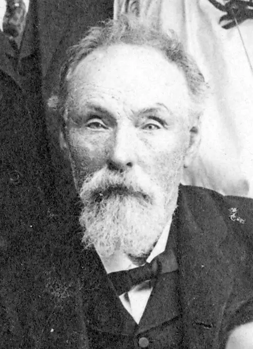 Black and white portrait of an old man with a silver mustache and long beard in a black suit with a black bow tie. He is looking straight at the camera with a serious look.
