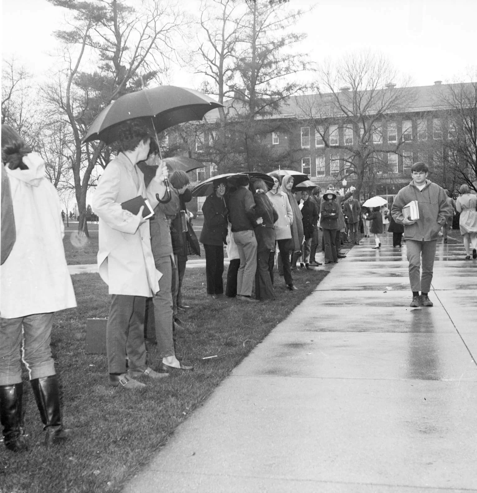 Black and white photo of people standing with umbrellas in the grass along a sidewalk.