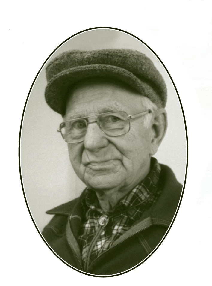 An older light-skinned man looks at the camera with maybe a bit of a judgmental look on his face. He's wearing a wool flat cap, flannel collard shirt, a bolo tie, and a jacket. He is wearing glasses.