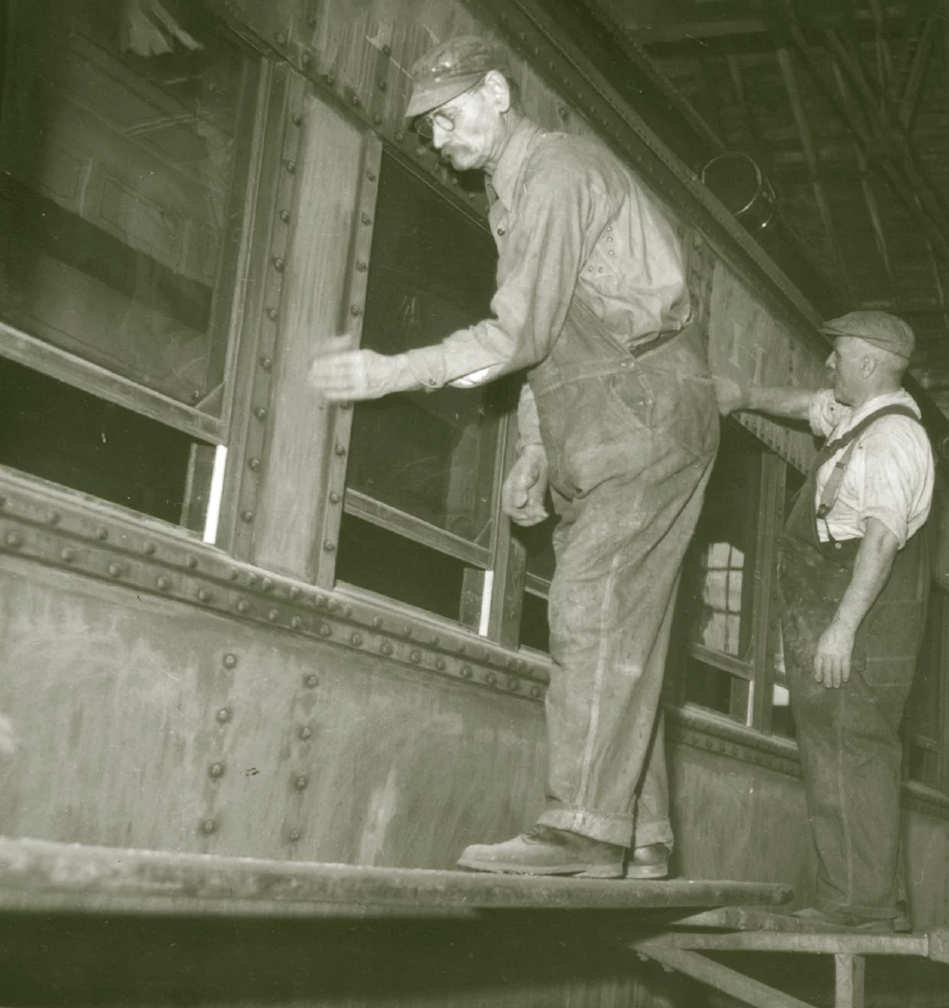 Two men are standing on scaffolding and are hand-sanding or wiping down the outdoor of a passenger car. They are both wearing hats and overalls, and have musaches.
