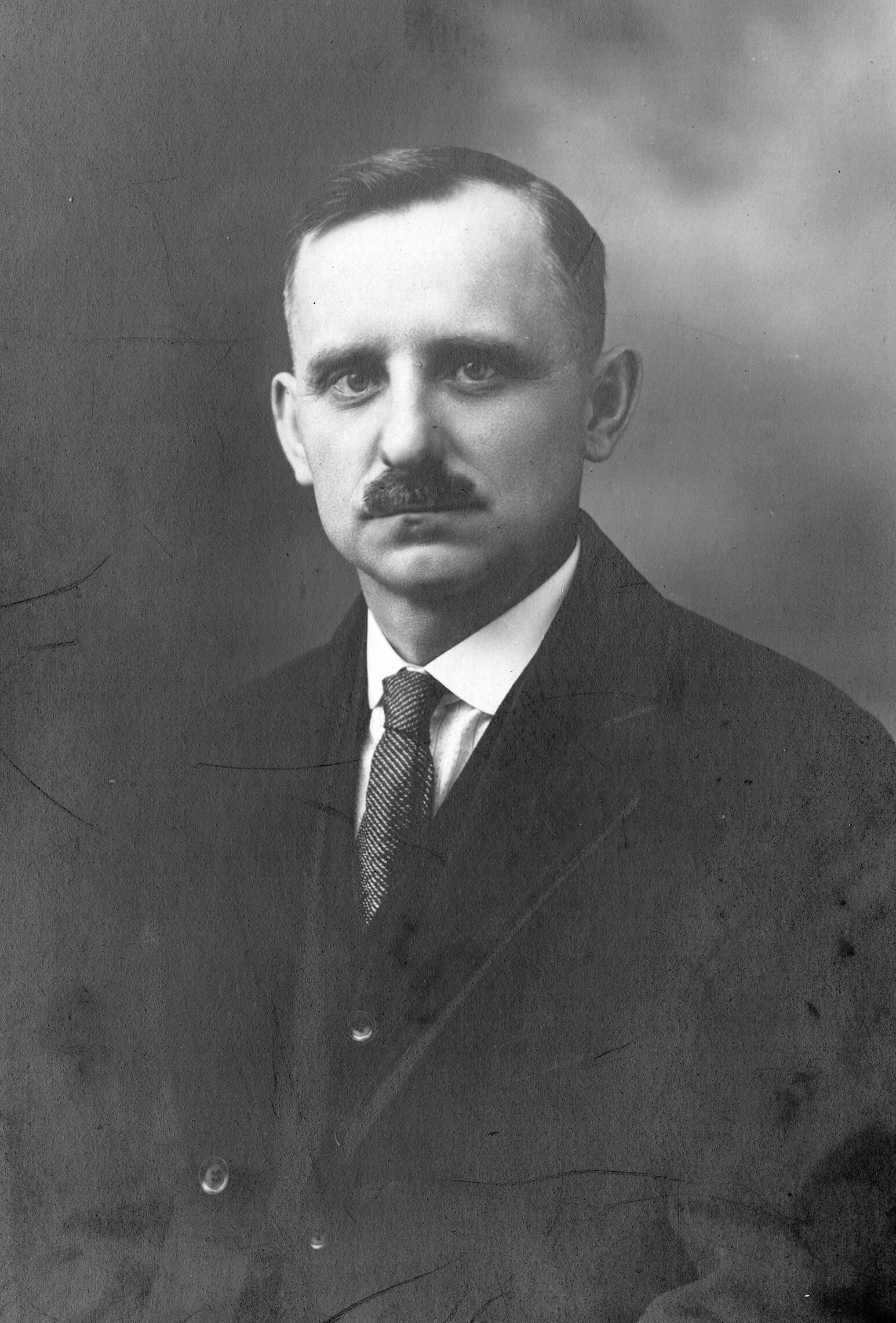 Black and white portrait of a young man in a suit, with thick mustache. He is staring into the camera with a serious face.