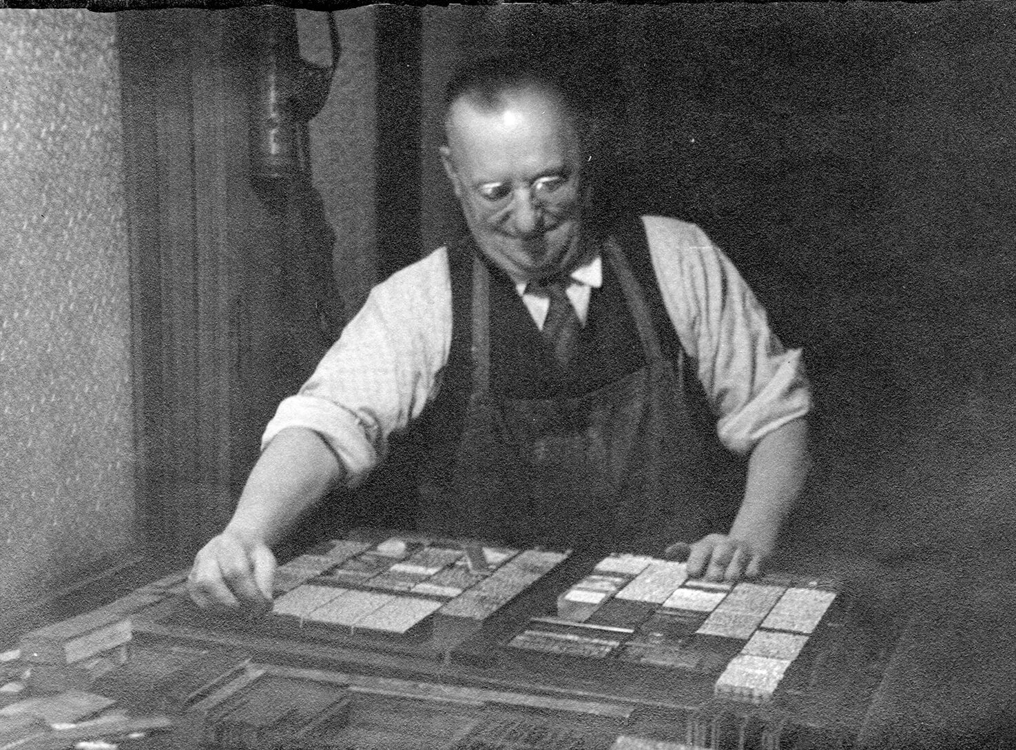 Black and white photo of a man wearing glasses and an apron, setting the type in a printing press