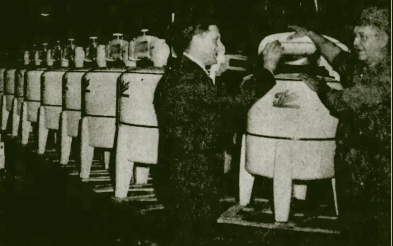 Two men stand in the foreground smiling and looking at a white washing machine. Machines are lined up as far as the eye can see. The machines are large white drums with 4 legs.