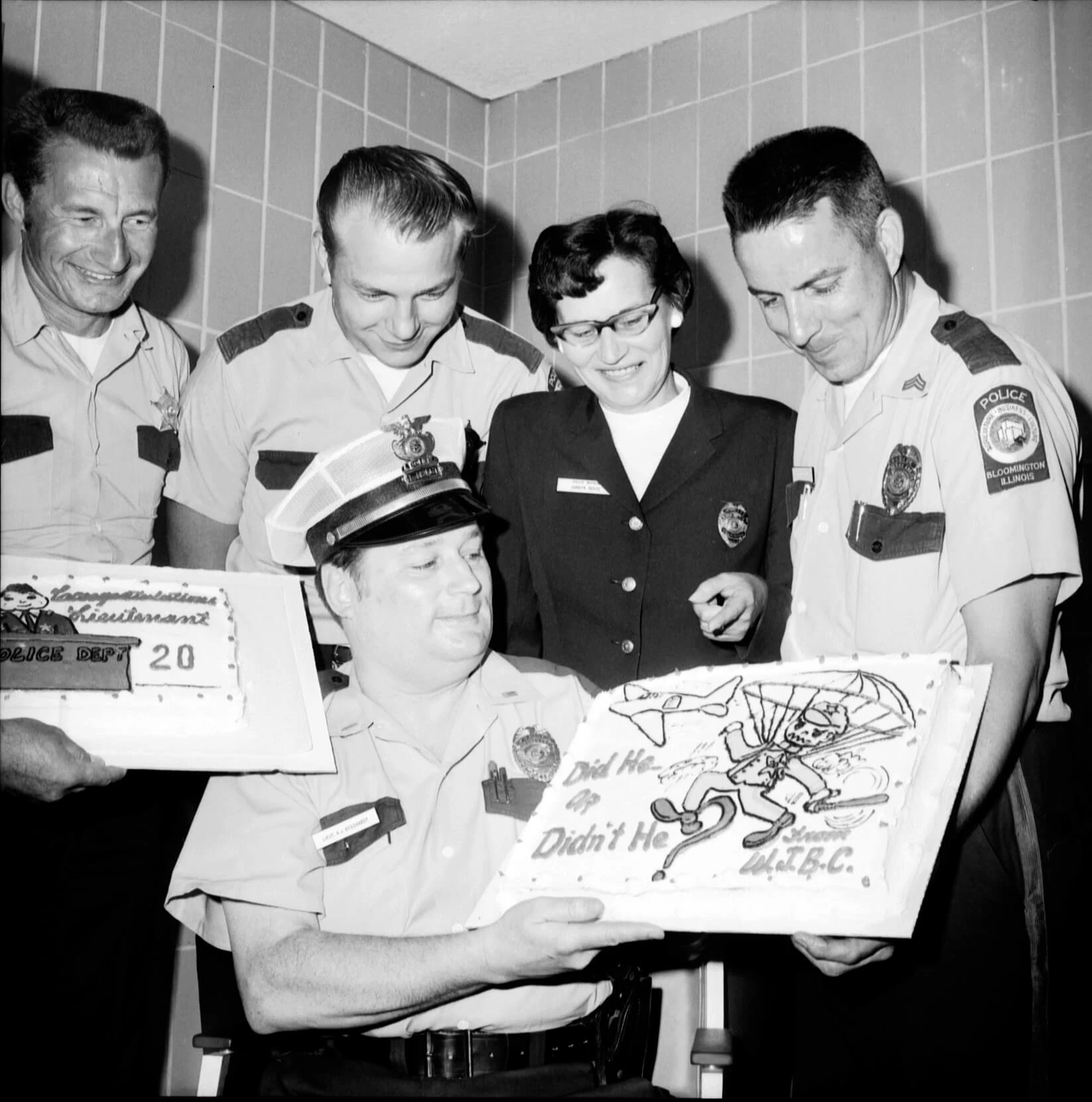 Four men in light-colored police uniforms pose with a woman in a dark police coat. They are holding two sheet cakes celebrating Reece's retirement. One cake shows a man on a parachute with a baton.