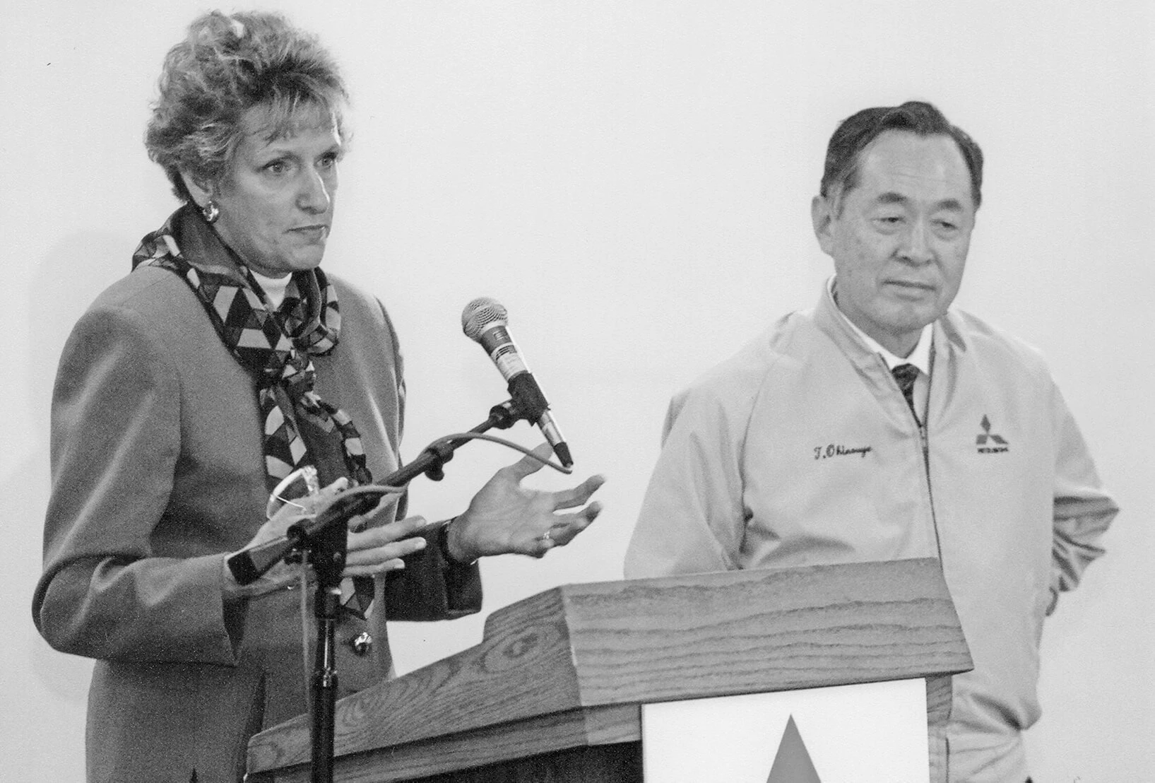 A white woman wearing a neck scarf, earrings, and short hair speaks at a podium. Next to her is a Japanese man wearing a Mitsubishi zip-up jacket, white shirt and tie.