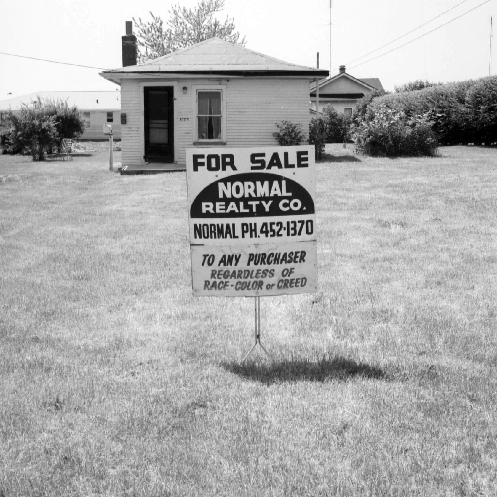 A sign in front of a small white house reads 'For Sale. Normal Realty Co. Normal Ph. 452-1370. To any purchaser, regardless of race, color, or creed.
