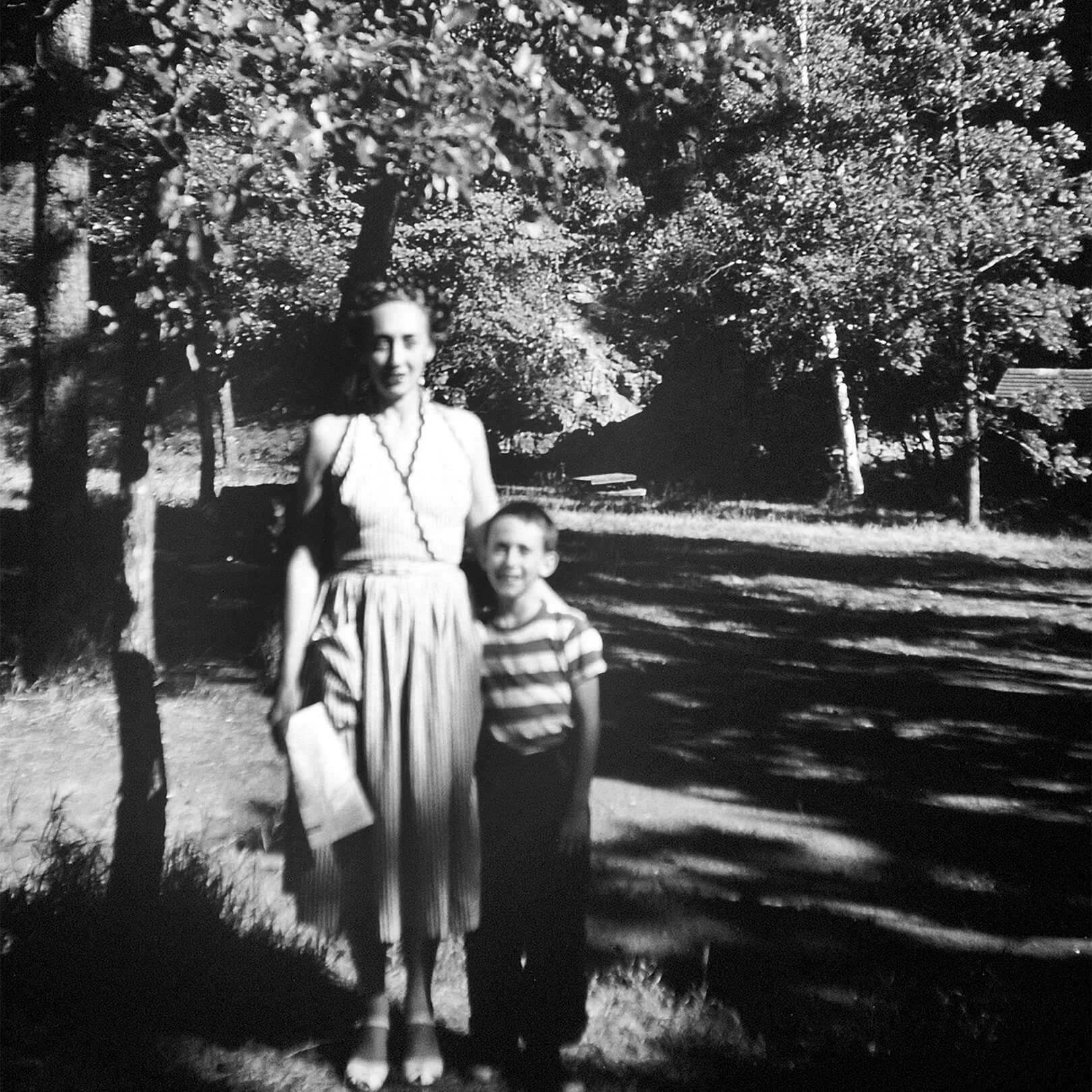 Black and white photo of a woman in a dress standing with a young boy.  They are standing outside in a yard.