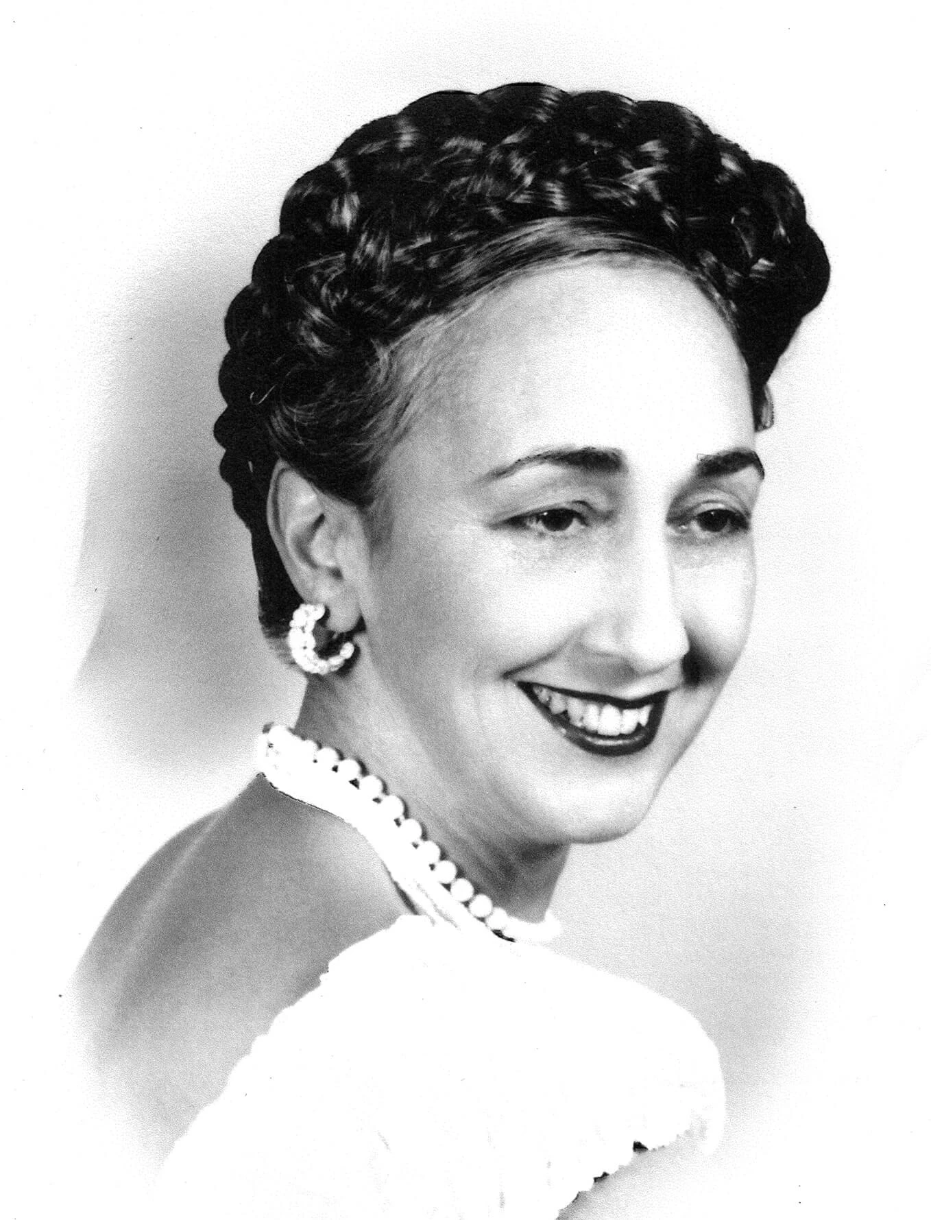 Lue Brown is wearing a white dress and pearls.  She has large earrings.  She is wearing red lipstick and she is smiling. Lue has dark eyes and dark hair. Her hair is braided and fashioned into an up-do on top of her head. She is facing the right and looking slightly over her shoulder.
