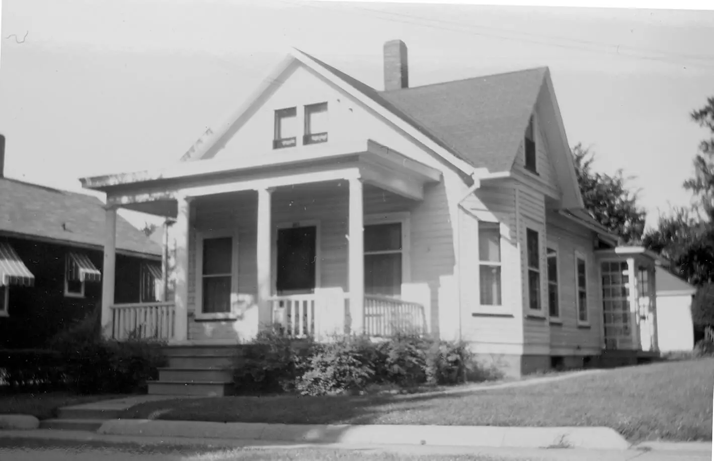 Black and white photo of a one-story home with front porch, white siding, and bushes in the front yard.