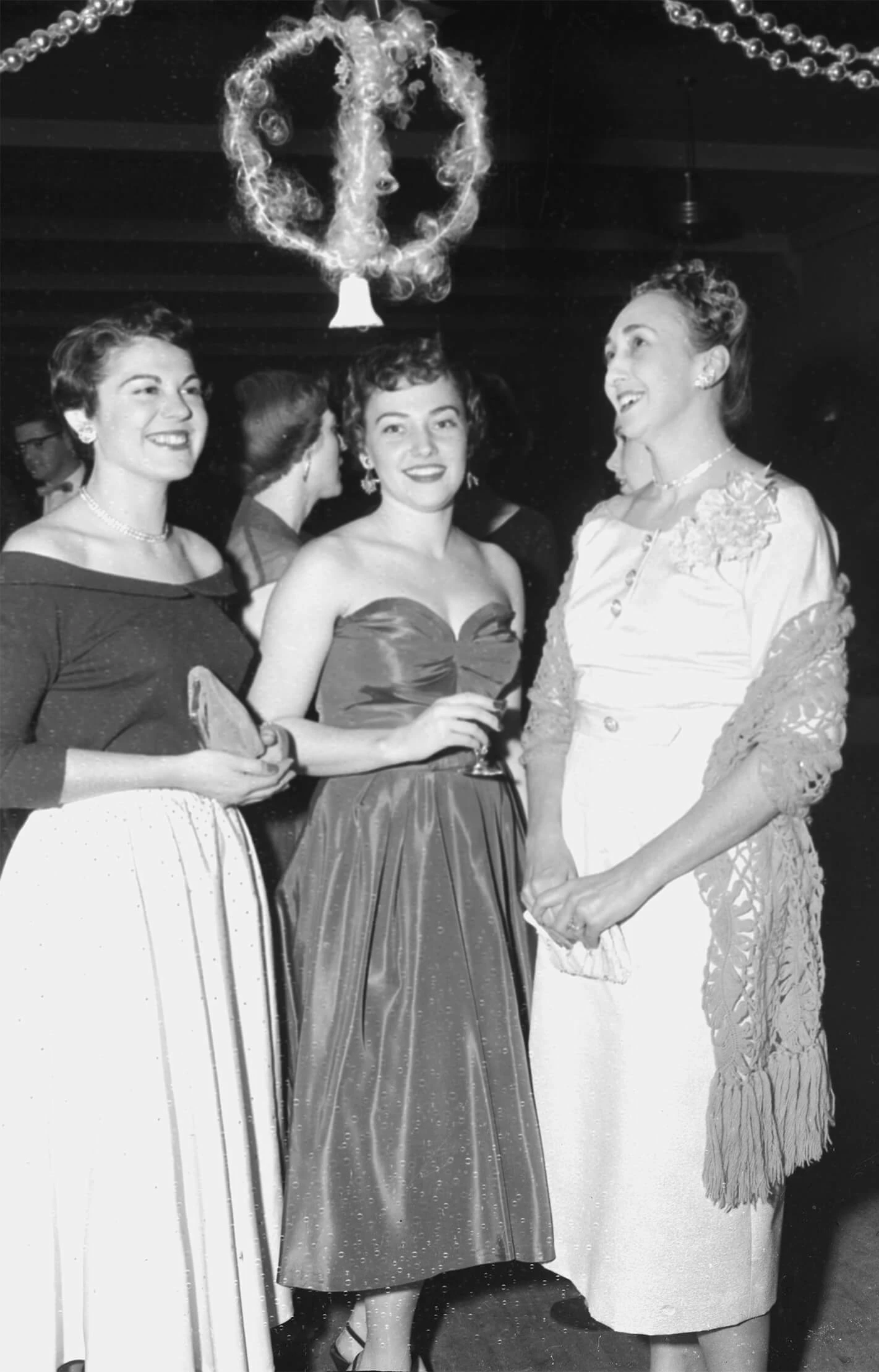 Black and white photo of three women in wearing formal dresses at a holiday party. One woman is carrying a glass and the other two women are carrying clutches.