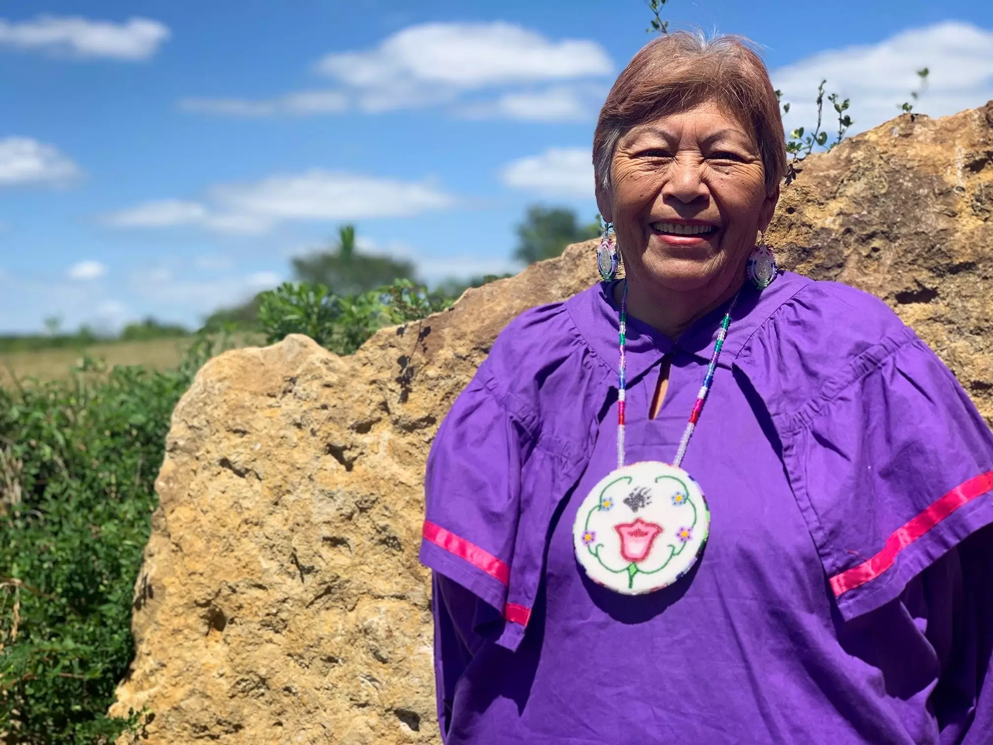 Color photograph of a Kickapoo woman with a warm, kind smile. She has short hair. She is wearing circle beaded earings and a circle beaded necklace. She is wearing a purple shirt with a pink ribbon detail on the sleeves. She is standing in front of a large boulder, and in the background you can see blue sky with white puffy clouds and a field that extend back to the horizon.