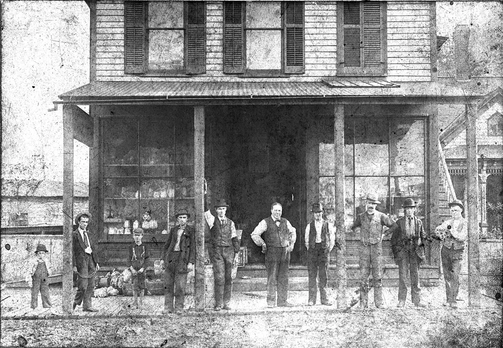 The O’Neil brothers, Daniel and William, are standing in front of their grocery store with their family. It is a two-story wood frame building with a covered front area.