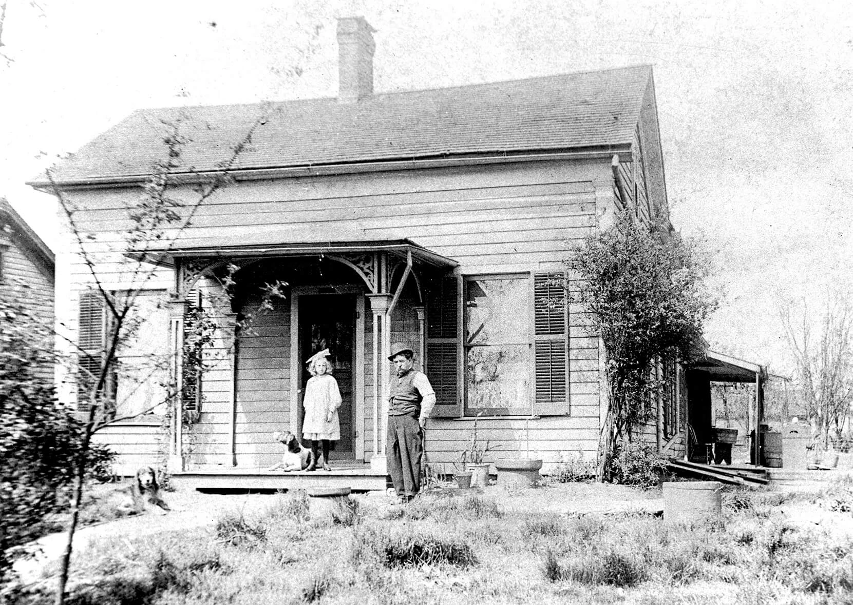 Black and white photo of a small, clapboard home with a front stoop. There is a little girl wearing a white dress standing on the stoop, with her dog at her feet. An older man in a top hat stands on the grass near her. There is a weedy lawn in front of the house.