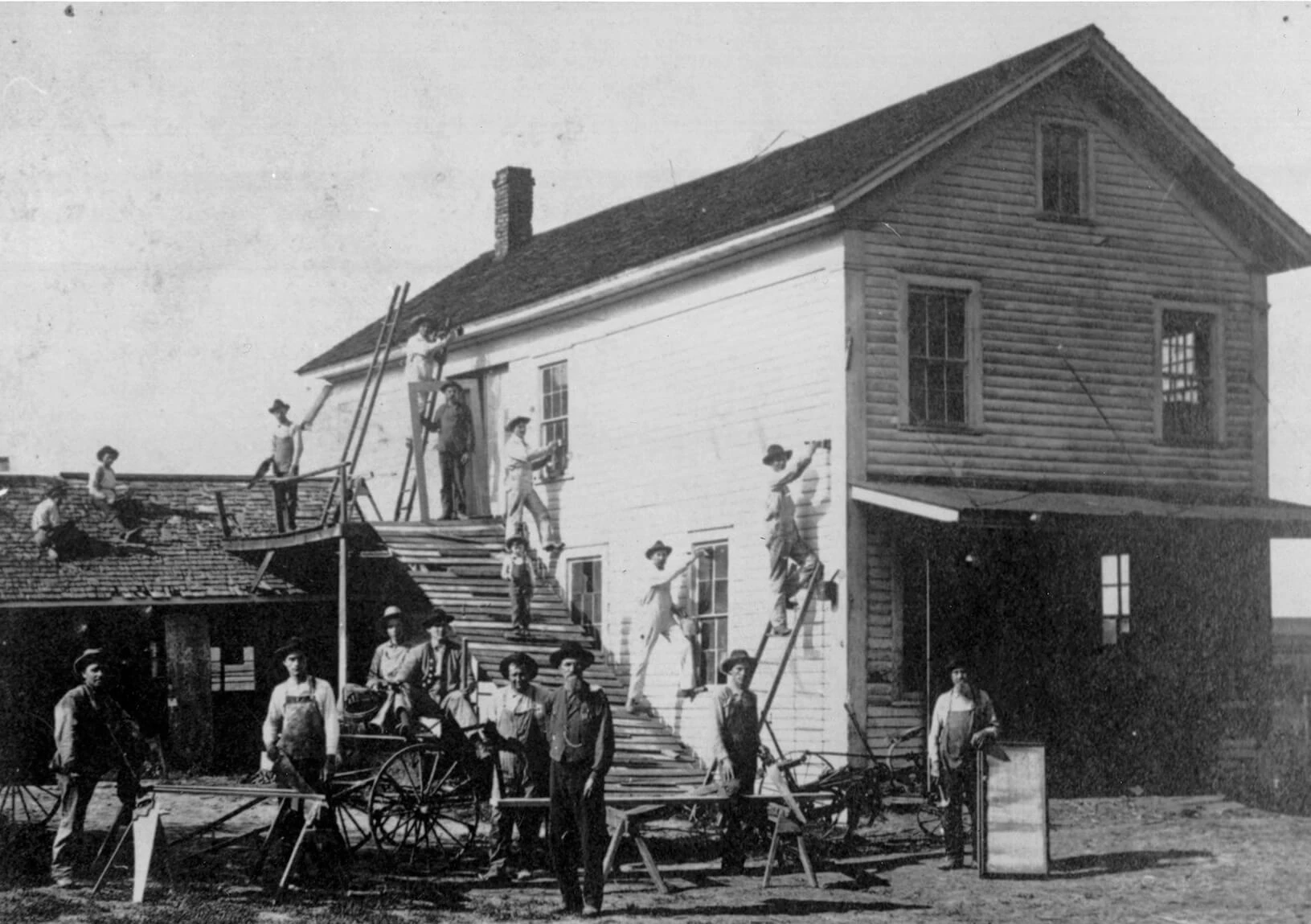 Black and white photo of many men working on a house. In the foreground men pose with their carpentry work and in the background men pose while painting and roofing.
